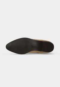 Semelle de chaussure beige montrant une semelle extérieure en caoutchouc noir texturé avec de fines rainures au talon et à l’avant-pied pour une bonne adhérence.
