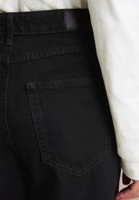 Detailný záber osoby v čiernych denimových nohaviciach s detailmi v zadnej vrecku a viditeľným rukávom bielej košele s dlhým rukávom.