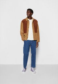 Brown and tan corduroy shirt jacket with buttons, paired with a cream t-shirt and blue denim pants. Footwear: purple high-top sneakers.