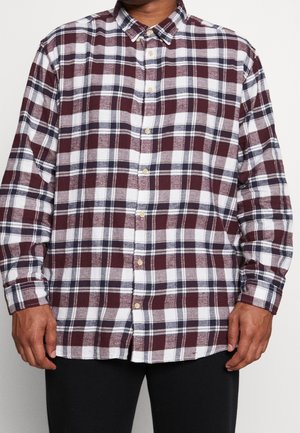 Mand iført en langærmet skjorte med knapper med et ternet mønster i bordeaux, hvid og navy samt sorte bukser.