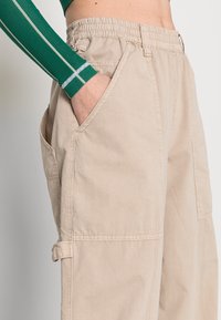 Beige cargobroek met een elastische tailleband, zijzakken en omgeslagen broekzomen. Het materiaal lijkt lichtgewicht met een gladde textuur.