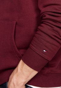 Burgundy sweatshirt med en framficka, ribbade ärmslut och en liten logodetalj på ärmen, som visar en mjuk, texturerad tyg.