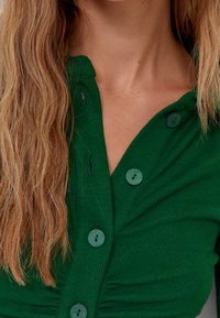 Haut vert côtelé avec un design à boutons, en tissu texturé, décolleté en V, boutons turquoise, souligné par des détails plissés.