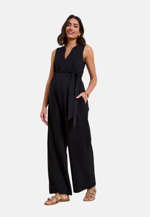 Smilende gravid kvinde iført sort ærmeløs jumpsuit med taljebånd og beige sandaler, stående med den ene hånd i lommen.