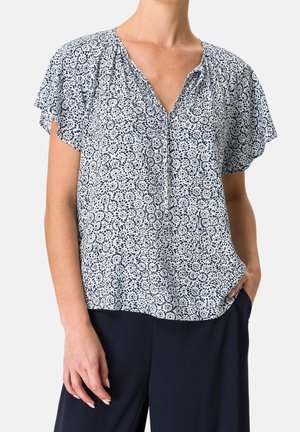 Woman wearing a short-sleeve white blouse with blue floral pattern and navy blue pants, one hand in pocket, against plain background.