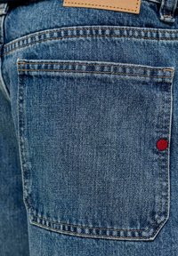Πίσω τσέπη από μπλε τζιν με ορατή πορτοκαλί ραφή και μια μικρή κόκκινη στρογγυλή ετικέτα στη δεξιά πλευρά.