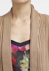 Gilet marron à côtes, design à l'avant ouvert, tissu texturé, superposé sur un haut à imprimé floral coloré avec un col en dentelle noire.