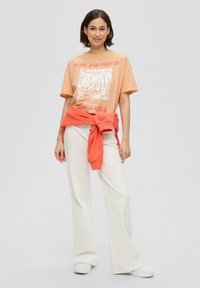 Pfirsichfarbenes T-Shirt mit weißem Grafikdruck, kombiniert mit lockeren weißen Hosen. Ein orangefarbener Pullover ist um die Taille gebunden. Lässiger Look.