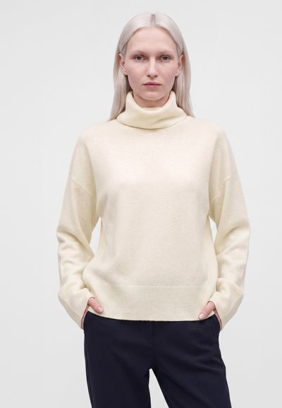 Femme aux cheveux blonds clairs et raides portant un pull à col roulé crème et un pantalon foncé, les mains dans les poches, sur un fond uni.