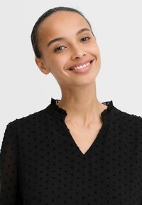 Blouse noire avec un motif texturé, dotée d'un décolleté en V et d'un col volanté. Tissu léger avec de petits points surélevés.