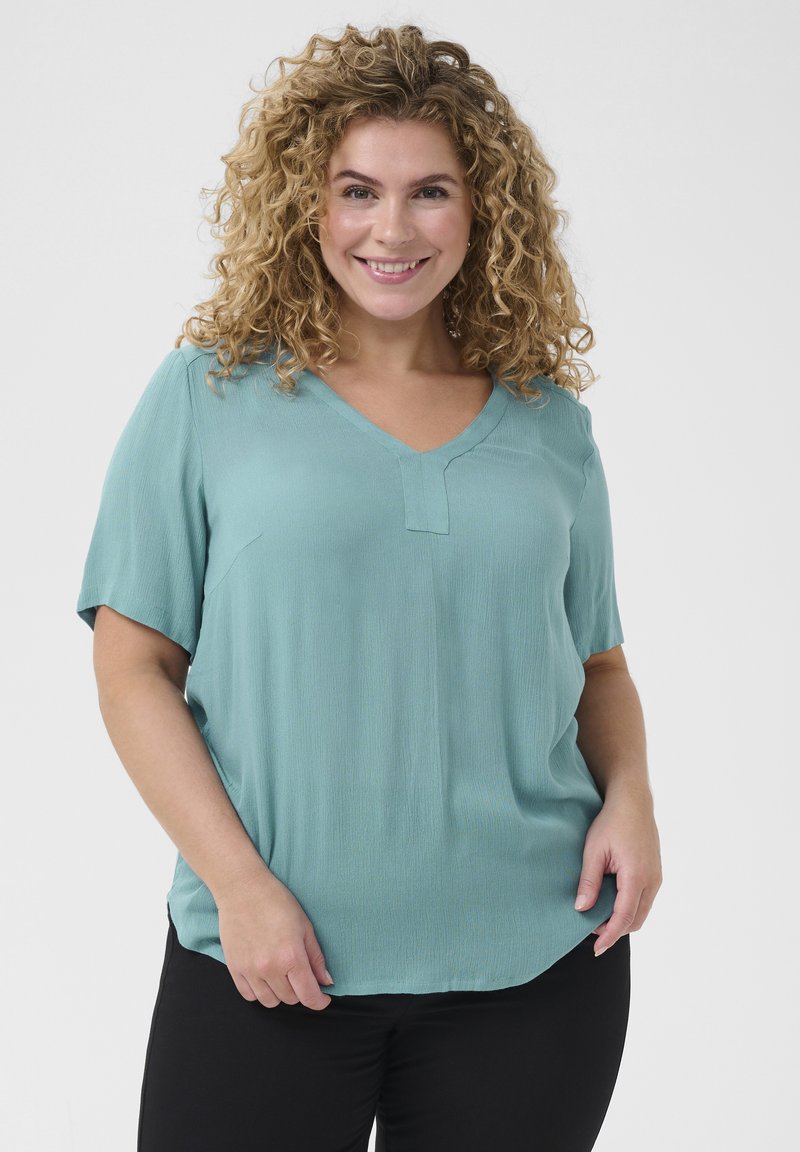Top corto teal con scollo a V, caratterizzato da una leggera texture e una vestibilità ampia, abbinato a pantaloni neri. Design semplice e versatile.