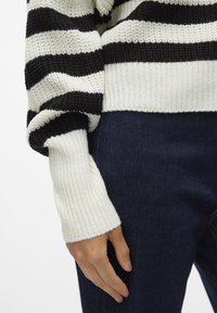 Black and white striped knitted sweater with ribbed cuffs, worn over dark denim jeans. The texture is chunky and the fit is relaxed.