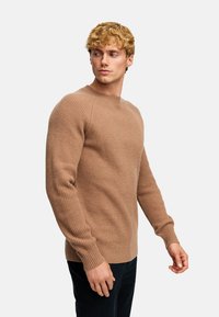 Jasno brązowy sweter w prążki z długimi rękawami, okrągłym dekoltem i teksturowanym materiałem, w połączeniu z ciemnoniebieskimi spodniami.