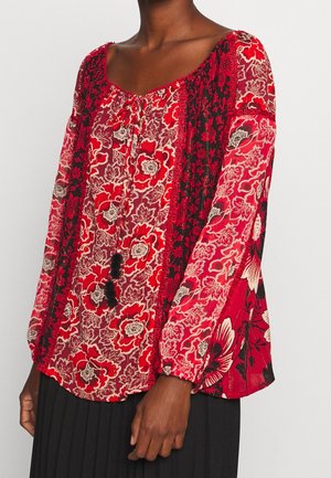 Femme portant un chemisier à manches longues rouge à fleurs avec un lien noir à pompons et des détails en dentelle complexe, associé à une jupe plissée noire.