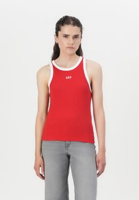 Rotes geripptes Tanktop mit weißem Rand und "GAP"-Logo auf der Vorderseite. Das Model trägt graue weit geschnittene Jeans. Schlichter Hintergrund.