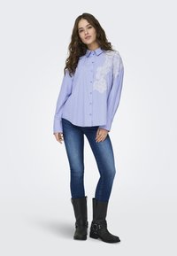 Camisa listrada azul clara e branca com detalhes em renda floral em um ombro, design abotoado, combinada com jeans azuis e botas pretas até ao joelho.