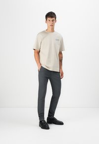 Beige Baumwoll-T-Shirt mit kurzen Ärmeln und einem bedruckten Logo auf der linken Brust, kombiniert mit dunkelgrauen taillierten Hosen. Weiche Textur, lässige Passform.
