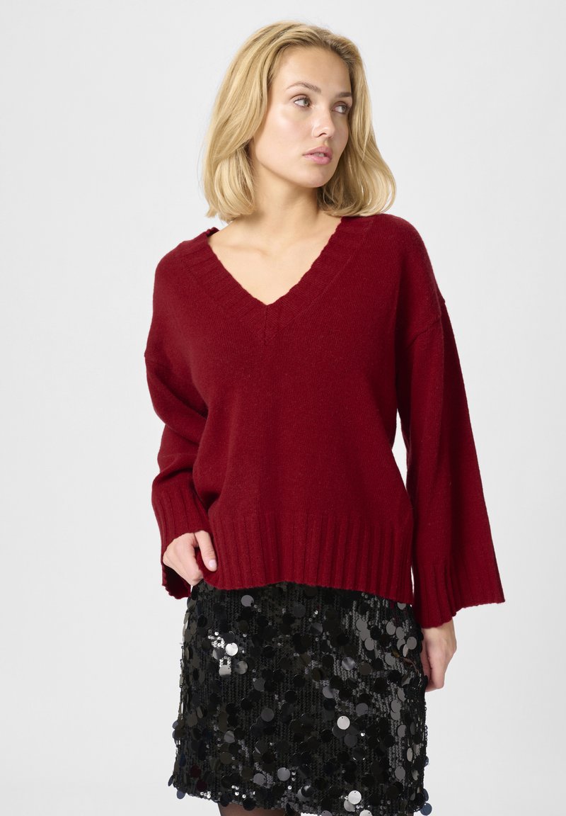 Bordeaux v-hals strikket sweater med ribbet kant og brede ærmer, parret med en sort mini-nederdel med pailletter og runde refleks accenter.