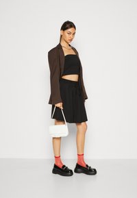Svart crop-top, svart skjørt med snor, brun blazer, hvit veske, rød sokker, og robuste svarte sko. Enkel bakgrunn.