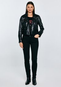 Nő áll előre nézve, fekete bőrdzsekit, fekete nadrágot, fekete bokacsizmát és fekete mintás pólót visel.