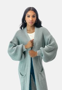 Lång, öppen framskjorta i ljusgrått, stickad struktur med ballongärmar och två framfickor, bärs över en vit topp.