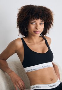 Donna con capelli ricci che indossa un reggiseno sportivo nero di Calvin Klein e mutande, appoggiata a una sedia beige contro uno sfondo bianco.