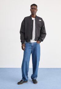 Giacca bomber nera con zip, polsini a costine e tasche laterali, indossata sopra una t-shirt bianca e abbinata a jeans blu larghi e scarpe nere.