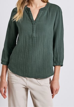 Vrouw draagt een donkergroene, getextureerde blouse met lange mouwen en een gespleten halslijn en beige broek tegen een effen achtergrond.