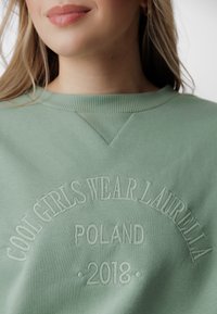 Felpa verde con colletto a costine e dettaglio a forma di V. Presenta un testo ricamato: "LE RAGAZZE FICHE INDOSSANO LAURELLA POLONIA 2018." Texture morbida.