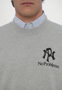 Sweatshirt gri cu guler dreaptă ribbată, prezentând un logo negru și textul "No Problemo" pe pieptul stâng, purtat peste o cămașă cu guler în dungi albastre.