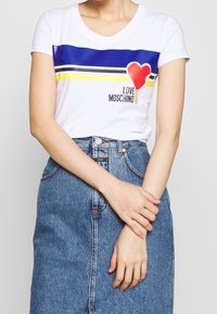 Bílý tričko s krátkým rukávem se modrými a žlutými vodorovnými pruhy, grafikou červeného srdce a textem "LOVE MOSCHINO". Nosit s džínovou sukní.