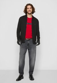 Veste zippée noire sur un t-shirt rouge avec logo, jeans gris et bottes en cuir noires jusqu'à la cheville. Les accessoires incluent des gants noirs.