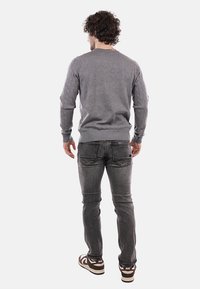 Maglione grigio con motivo a treccia sulle maniche, polsini e orlo a costine. Indossato con jeans grigi e scarpe da ginnastica scure, visto da dietro.