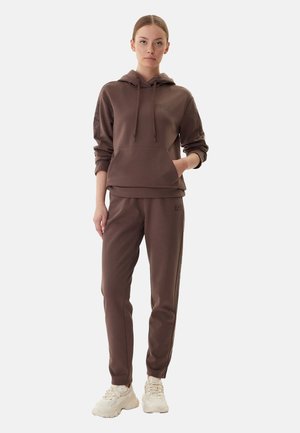 Donna che indossa una felpa marrone con cappuccio e pantaloni coordinati, con sneakers bianche chunky, in piedi contro uno sfondo chiaro e neutro.