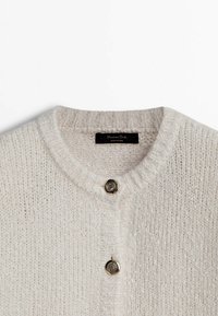 Lichtbeige gebreide cardigan met ribbeltextuur, voorzien van een ronde halslijn en twee decoratieve goudkleurige knopen aan de voorkant.