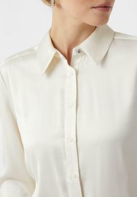 Cream blouse with a classic collar, front button closure, and a decorative metallic detail at the collar, featuring smooth, soft fabric texture.