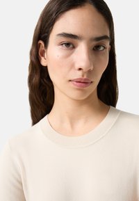 Strickpullover in Off-White mit Rundhalsausschnitt, geripptem Kragen und glatter Textur, die eng um die Schultern anliegt.