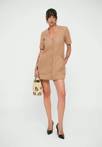 Kvinna iklädd en beiget knäppt kortärmad klänning med framfickor, hållande en vävd handväska och bärande svarta högklackade skor.