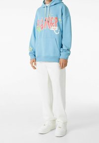 Persona vistiendo una sudadera ligera azul con texto gráfico y estrellas, pantalones blancos y zapatillas blancas, de pie contra un fondo blanco liso.