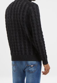 Pull noir en maille torsadée avec un motif texturé, poignets et ourlet côtelés ; associé à un jean bleu clair avec un accent décoratif sur la poche arrière.