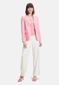 Lichtroze blazer met een getailleerde pasvorm, gelaagd over een gemusterde top en gecombineerd met vloeiende witte broeken en roze hakken.