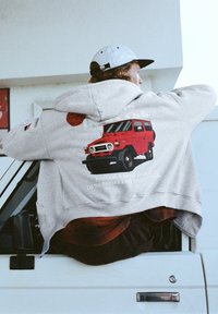 Šedá mikina s kapucí zdobená grafikou červeného Toyota Land Cruiser na zádech a textem pod ním. Nahoře nošena přes flanelovou košili, na pozadí vozidla.