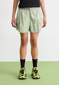 Pantalones cortos estilo cargo de color verde claro con cinturilla elástica, que cuentan con bolsillos laterales y una textura suave. Se llevan con calcetines negros y zapatillas amarillas.