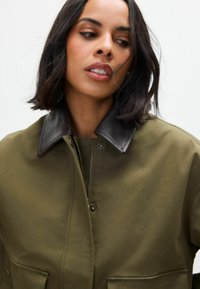 Olivgrüne Jacke mit braunem Lederkragen, Reißverschluss und zwei Fronttaschen. Glatte Textur mit strukturiertem Design.