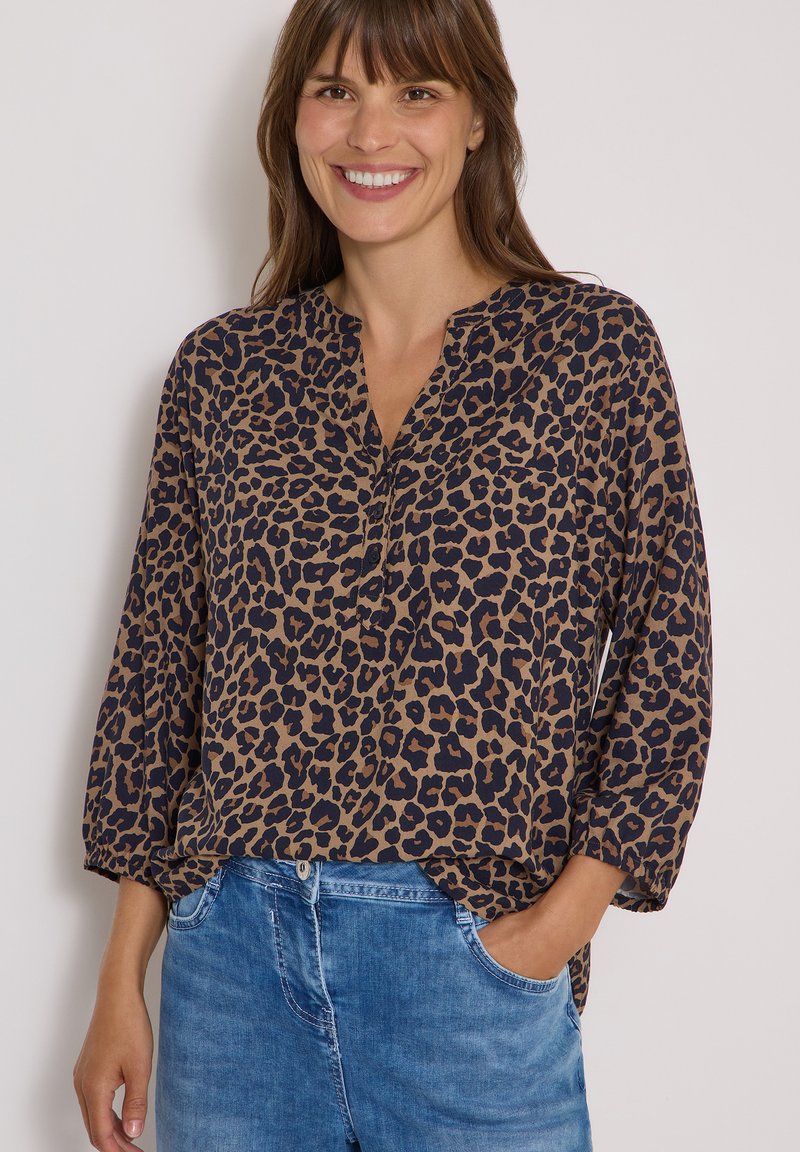 Femme souriante portant un chemisier marron et noir à imprimé léopard avec manches trois-quarts et un jean bleu, une main dans la poche.