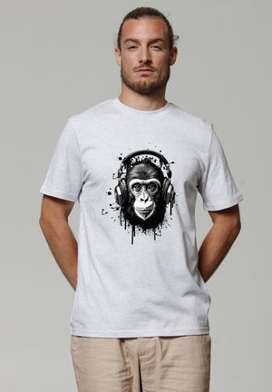 Man met naar achteren gebonden haar, draagt een lichtgrijs t-shirt met een zwart-wit chimpanseegezicht met koptelefoon en verfspatontwerp.