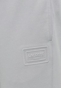 Ανοιχτό γκρι φόρμα από απαλό ύφασμα με υφασμένη ετικέτα λογότυπου που φέρει την ανάγλυφη επιγραφή "LACOSTE PARIS". Απλός σχεδιασμός, χωρίς ιδιαίτερα μοτίβα.