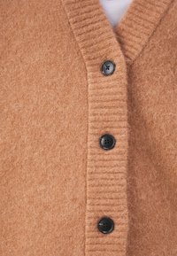 Cárdigan suave de punto marrón con escote en V acanalado y tres botones oscuros abrochados sobre una camiseta blanca.