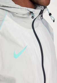 Veste légère grise avec capuche ; présente une fermeture éclair noire et des accents turquoise, y compris un logo Nike et des détails de couture.