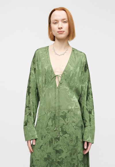 Πράσινο σατινέ φόρεμα με μακριά μανίκια, βαθύ V λαιμόκοψη, λεπτομέρεια με κορδόνι στο μπροστινό μέρος και διακριτό υφασμάτινο μοτίβο σε όλη την επιφάνεια. Ασημένιο κολιέ ως διακριτική λεπτομέρεια.